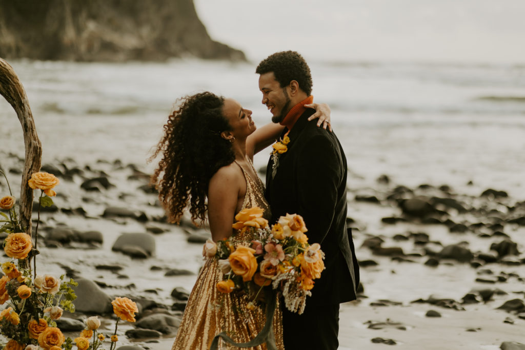Happy Wedding Couple with Golden wedding Dress and Orange Wedding Bouquet sharing vows on Short Sand Beach for their Oregon elopement ceremony. 