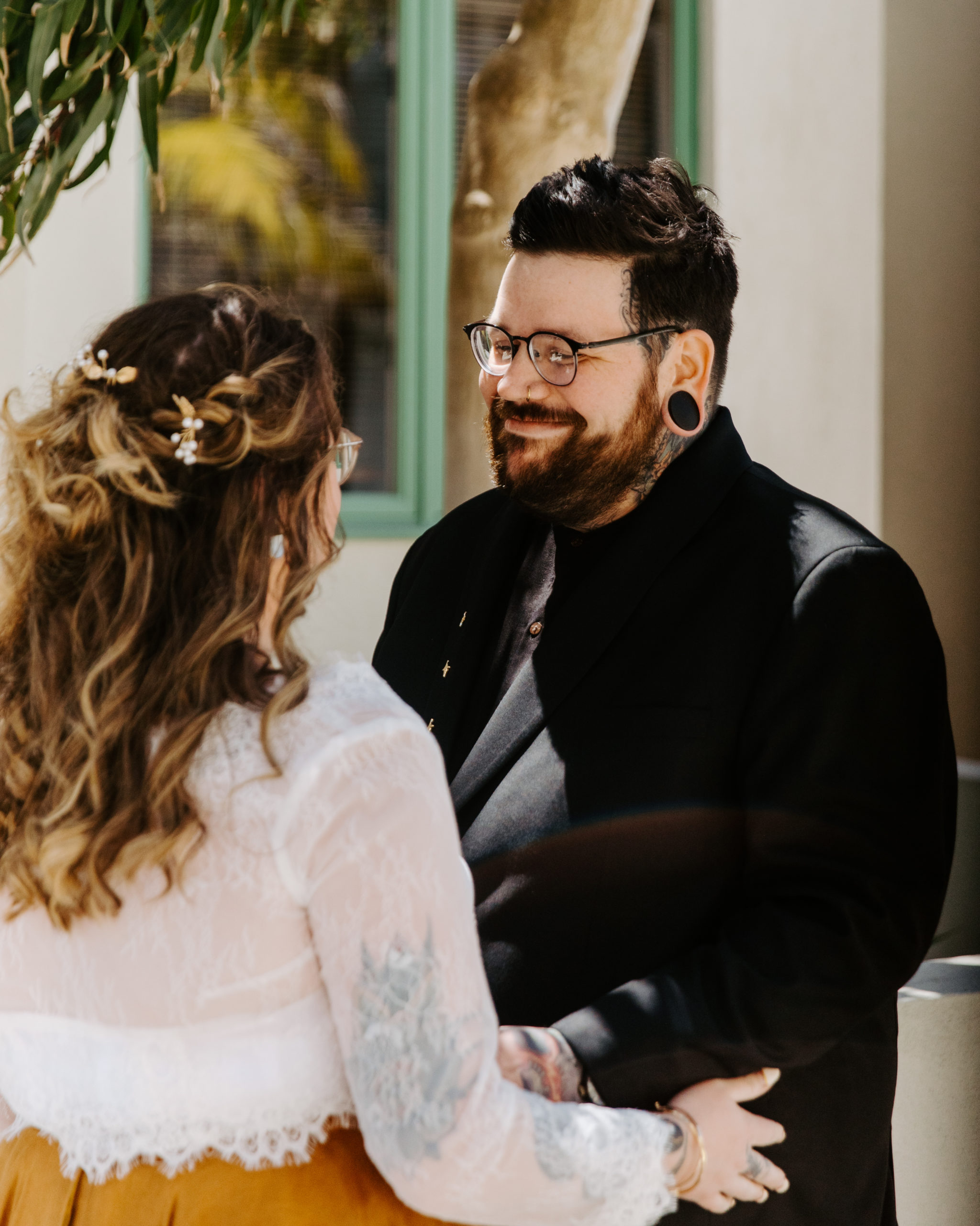 A happy couple at their wedding in San Diego California. The groom is in a black tux and the bride is in a white lace dress with orange skirt. Both are smiling, it is a sunny day. 