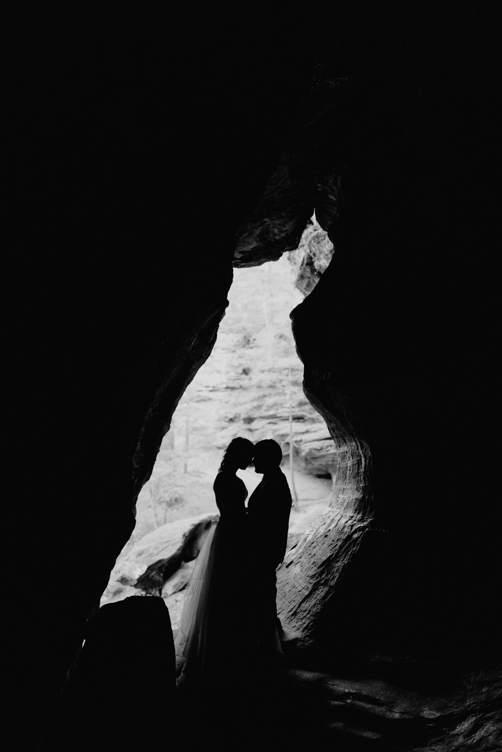 A hocking Hills Elopement couple posing in a cave entrance. The couples silhouette is filling the frame of the cave and the photo was taken in black & white