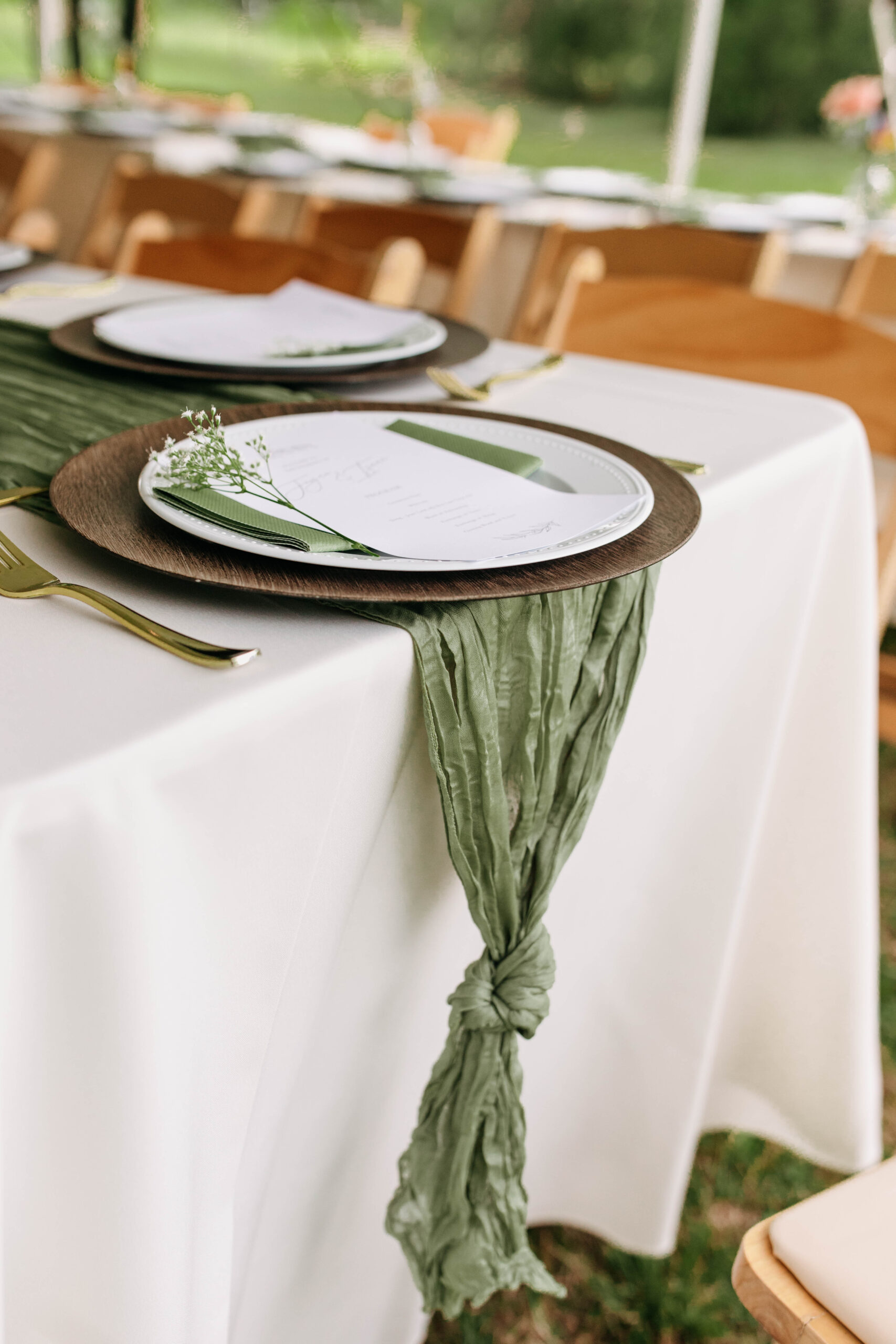 Photograph of green garden wedding reception decor. The wedding was an outdoor tent wedding with wooden chairs and white tablecloths. In this photo you can see the gold silverware, baby breath reception setting decor, and the wooden table chargers.