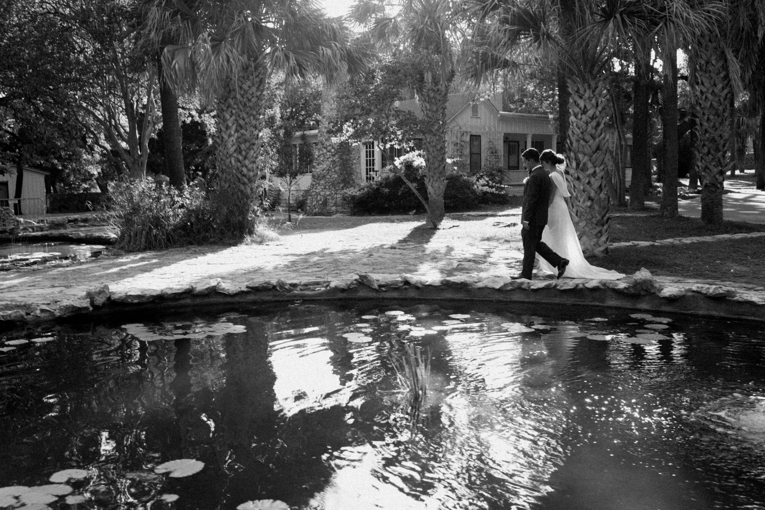 A black and white photograph of a wedding couple walking by the pond at Mayfield Gardens. 