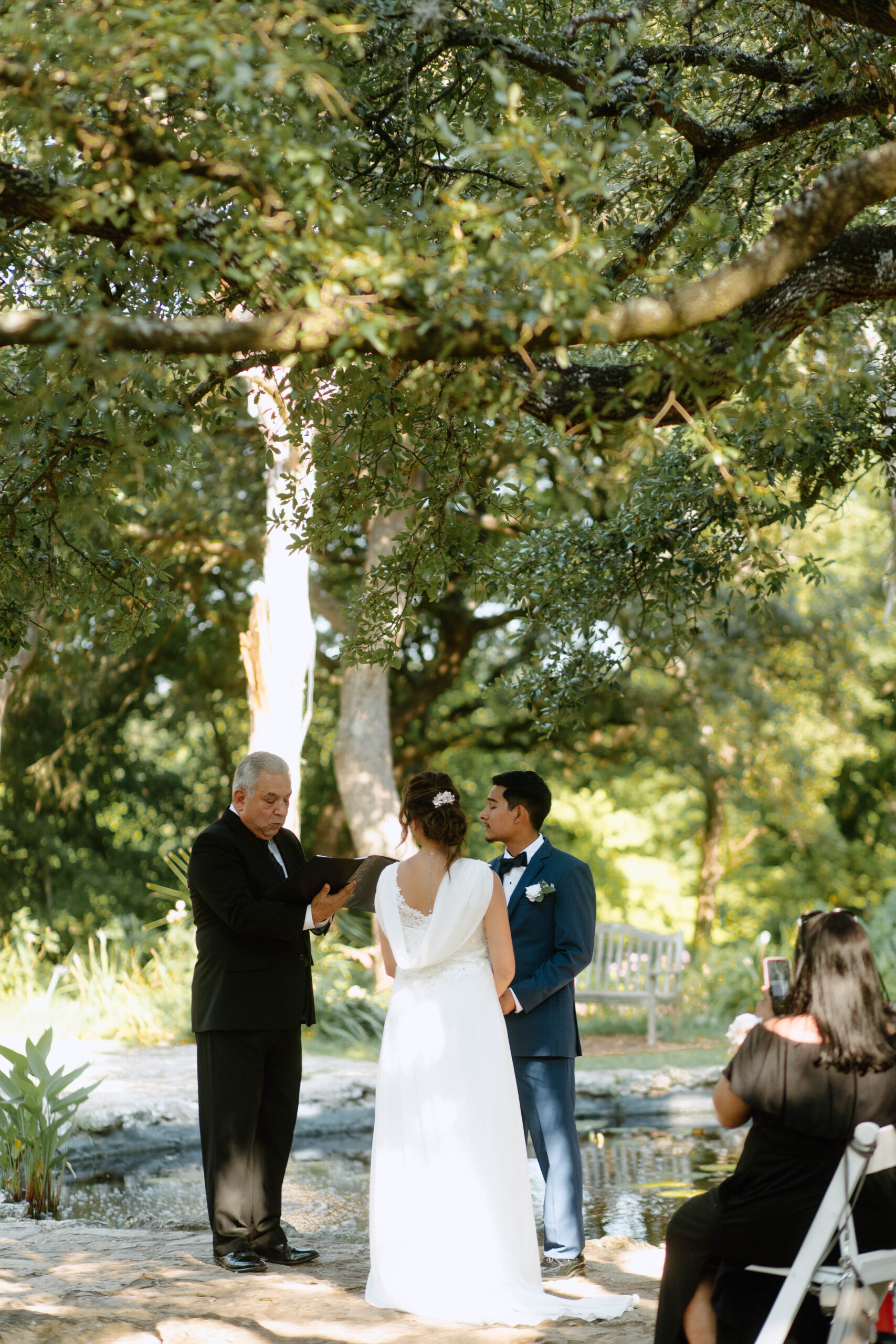 Photograph of a wedding ceremony at Mayfield Park. The wedding couple, Ana and Abel sharing vows next to a pond. 