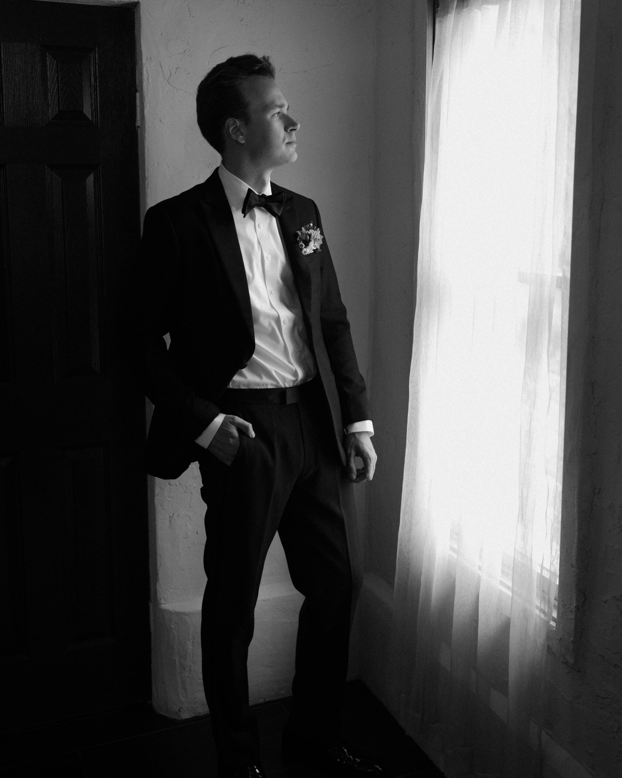 Black and white wedding photo of groom getting ready on his wedding day. The groom is wearing a classic black suit, white shirt and black bowtie.